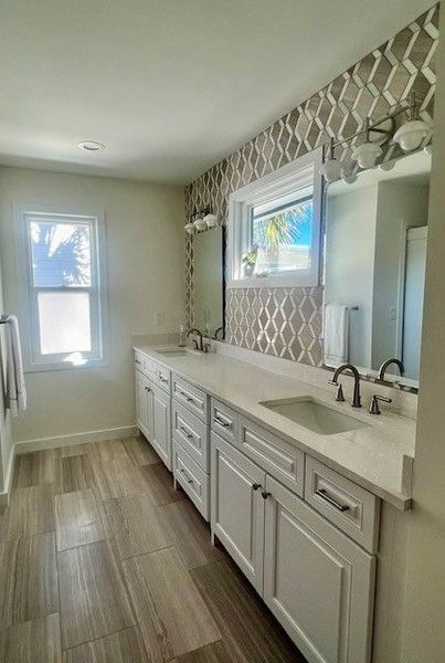 Bathroom with white vanity, patterned backsplash, two sinks, and wood-look tile floor.