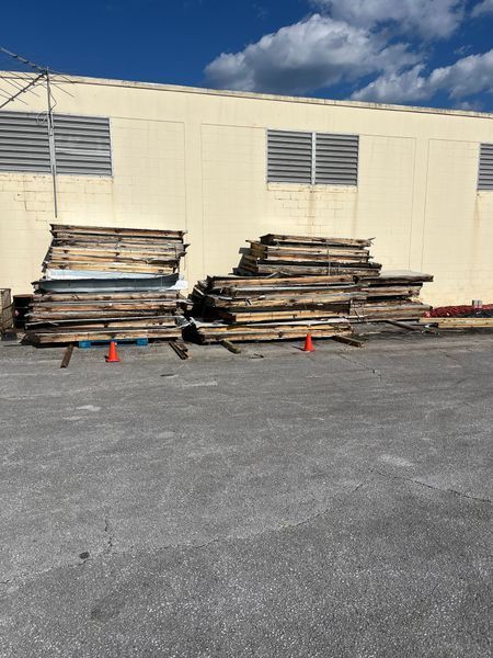 Piles of weathered wooden planks rest outside a tan building, with two orange traffic cones on the pavement.