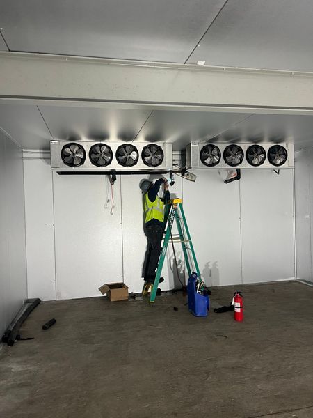 A person on a ladder is installing refrigeration units in a white-walled room.