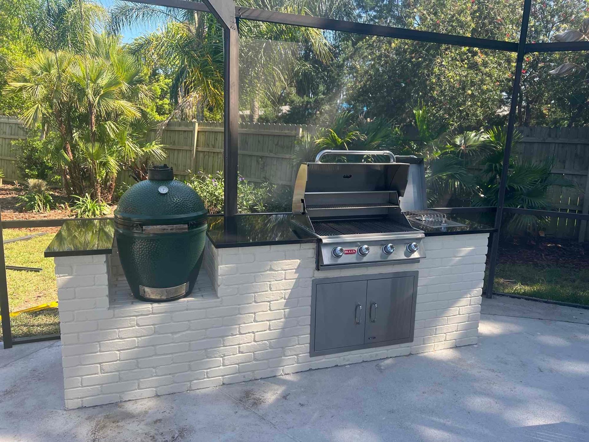 Outdoor grilling station with a green egg smoker, gas grill, and stainless steel cabinet on a white brick structure.