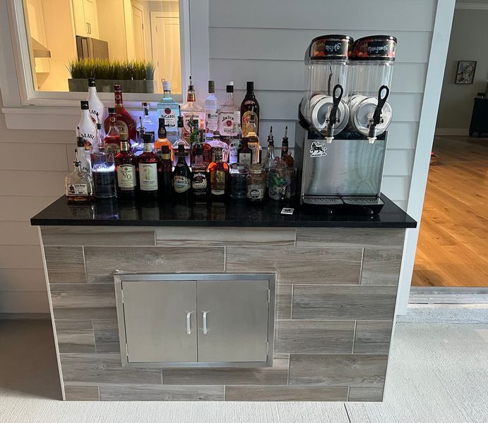 Outdoor bar with liquor bottles, two-tank slushy machine, and a stainless steel storage cabinet.