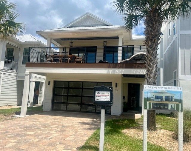 Two-story beach house with light siding, a balcony, and a garage; a sign in the yard.