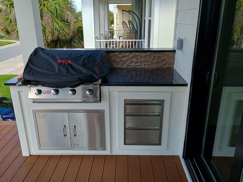 Outdoor kitchen with grill, cabinet, and countertop on a deck. Covered grill, stainless steel features, and textured backsplash.