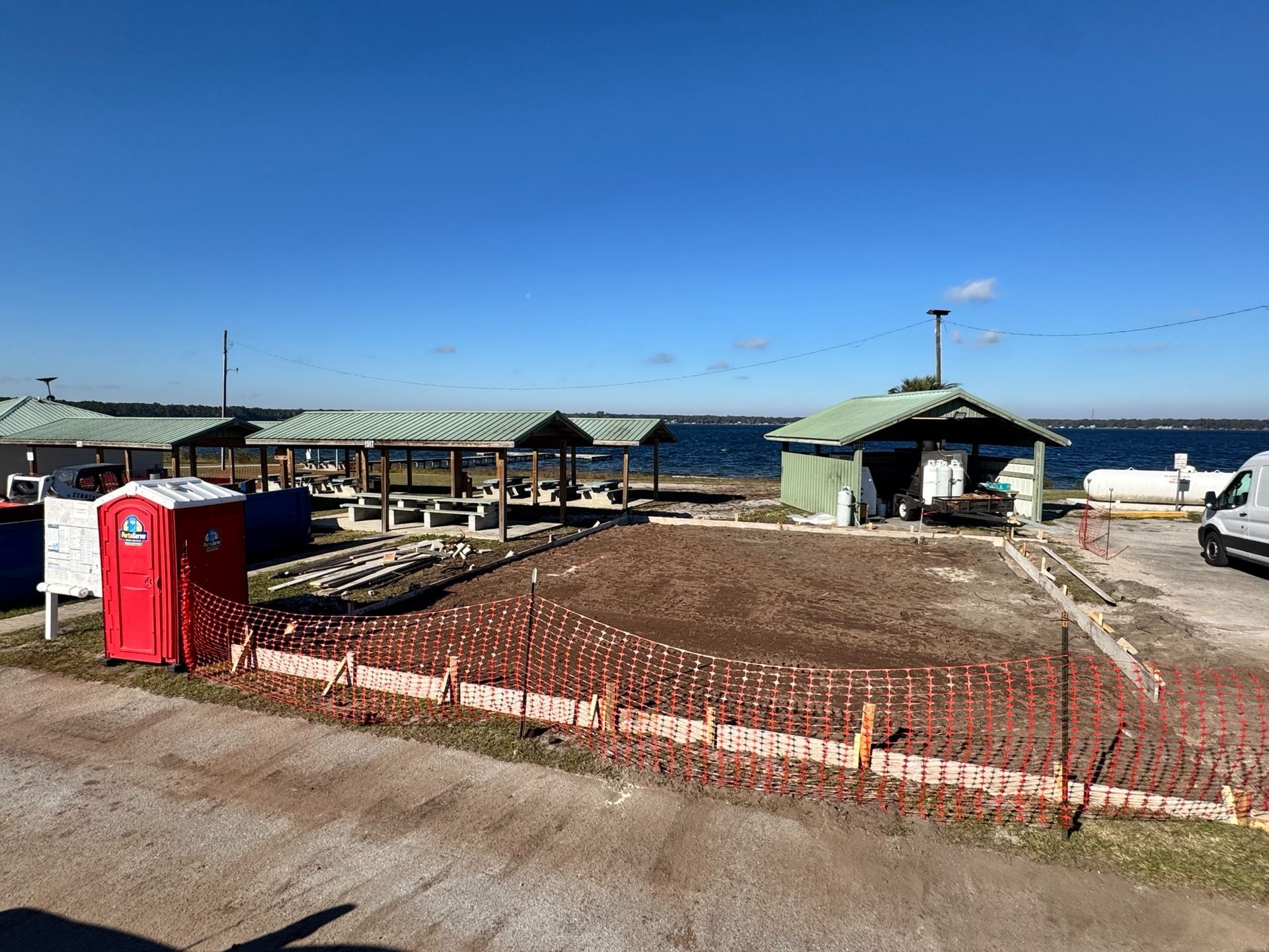 Construction site by water with covered picnic tables, small building, red porta-potty, and blue sky.