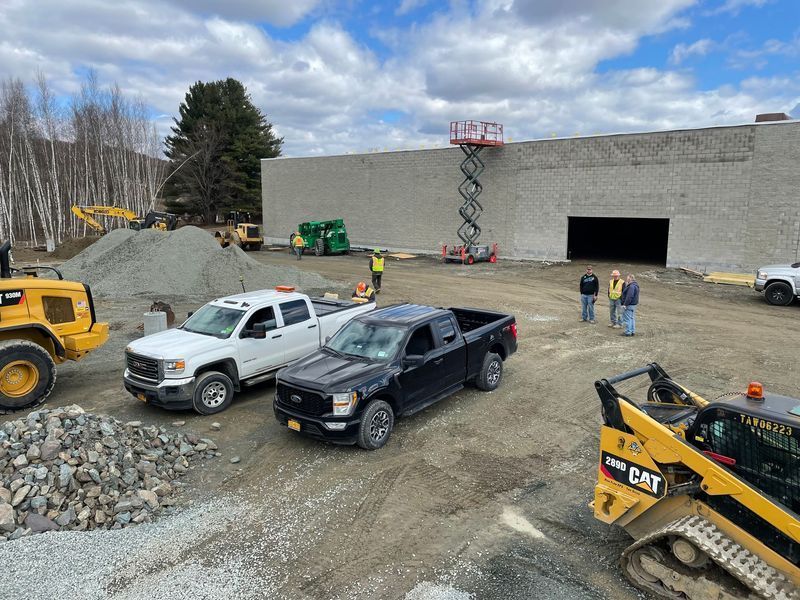 Construction of a tan-walled building with a steel frame, in a grassy field with trees.