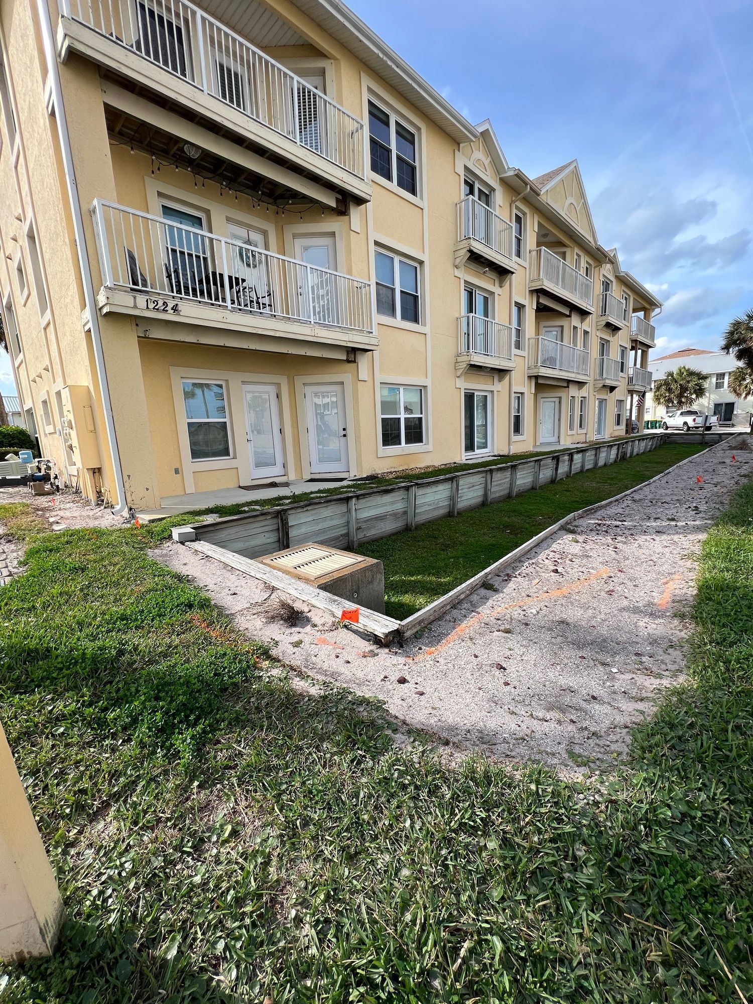 Yellow multi-story building with balconies, alongside a grassy area and a gravel path.