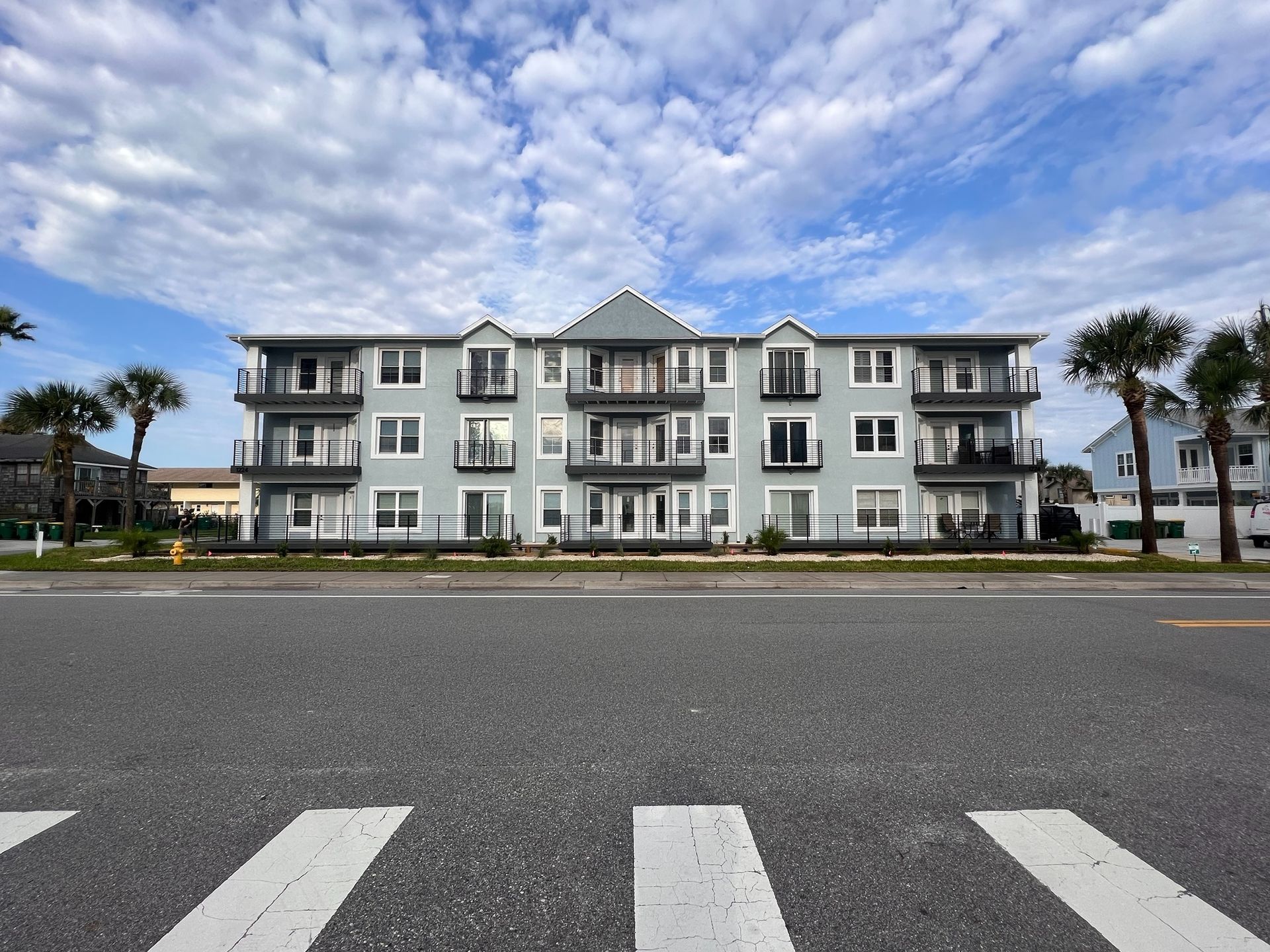 Light blue apartment building with balconies, set against a cloudy sky. Street in foreground.
