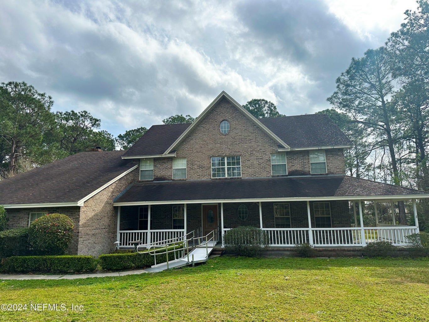 Brick house with wrap-around porch and ramp, set in a grassy yard under a cloudy sky.