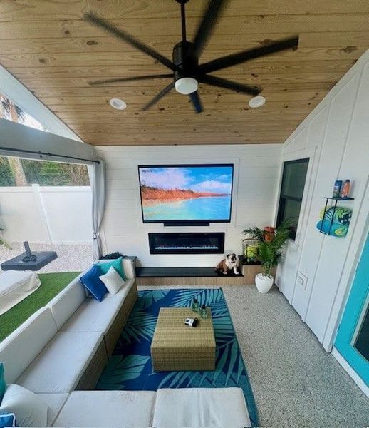 Outdoor living space with TV, fireplace, sectional sofa, ceiling fan, and blue accents.