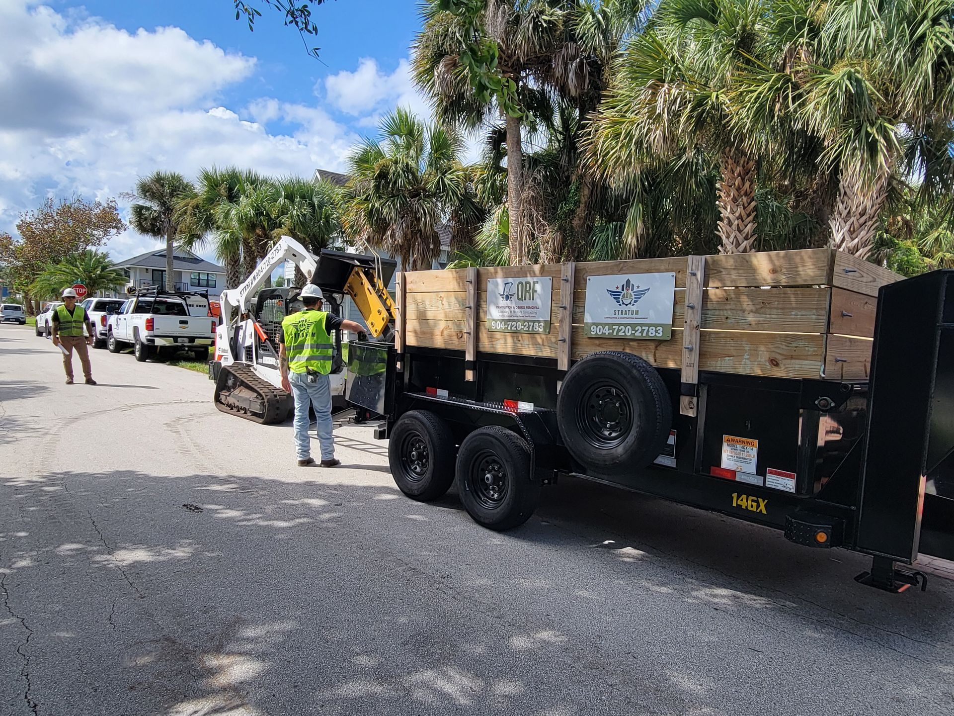 Workers loading wooden planks onto a trailer with an excavator on a residential street.