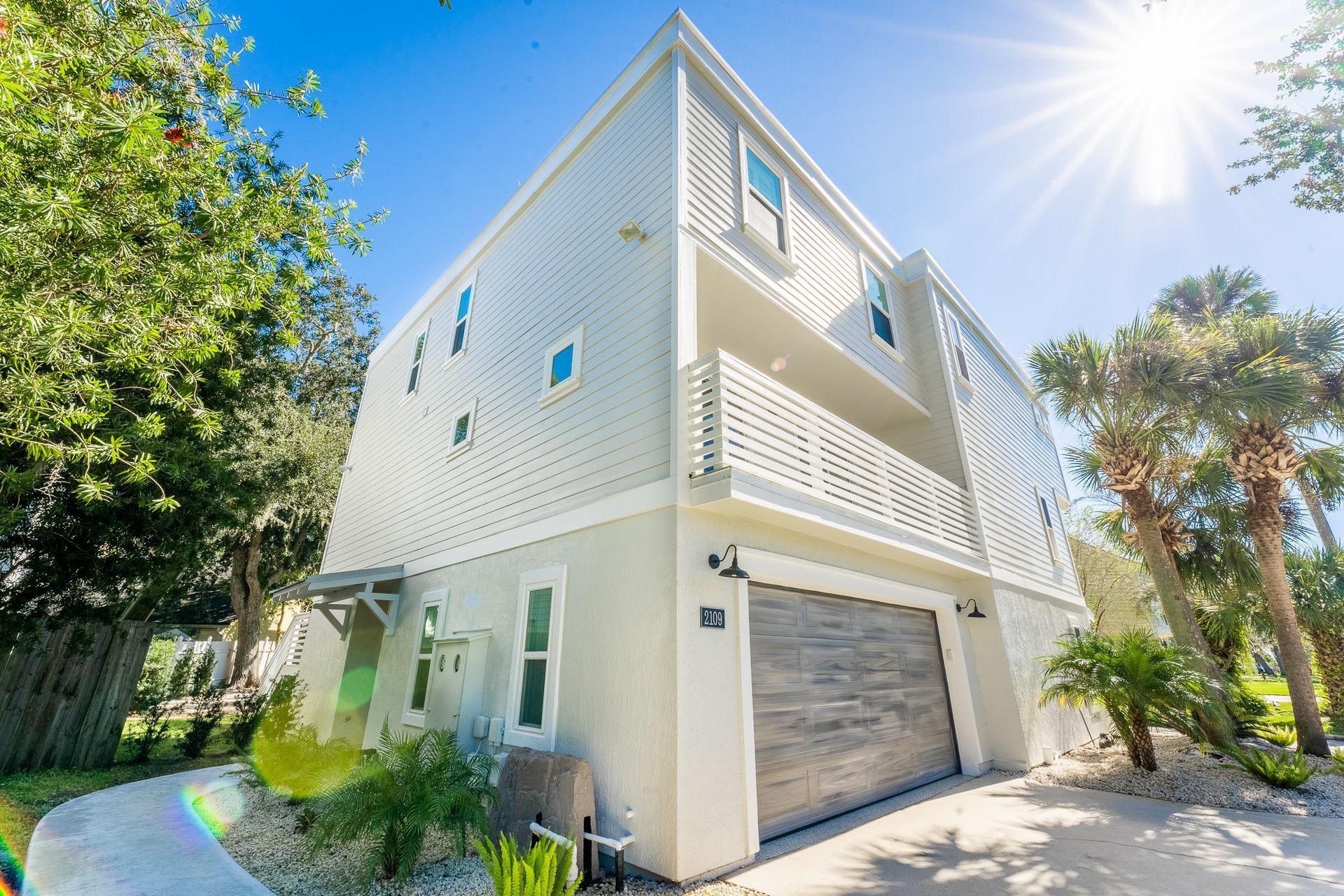 Modern multi-story house with gray garage door, white siding, and balcony, under a bright sun.