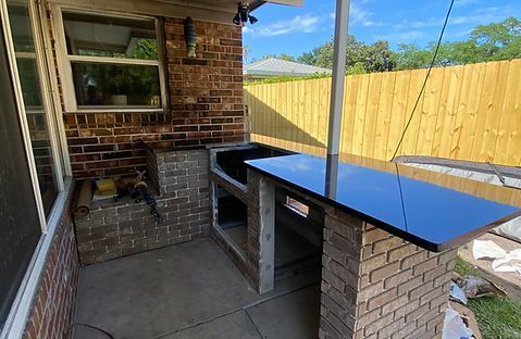 Outdoor brick bar with black countertop, against a brick wall, with a wooden fence in the background.