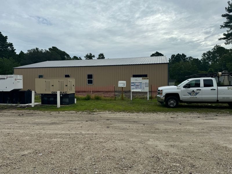 A brown building with a metal roof, generators, a truck, and a sign on a gravel lot.
