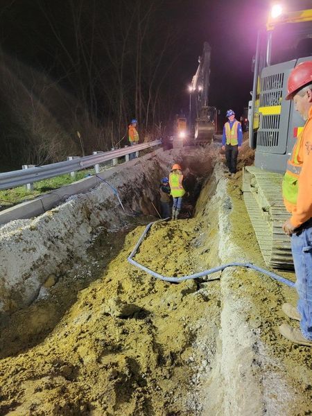 Construction workers in reflective vests working at night in a long trench next to a road.