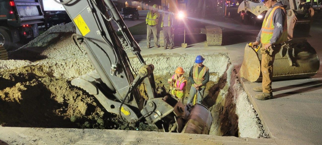 Workers and excavator at a construction site digging a trench in the road at night.
