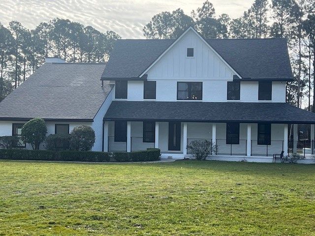 White two-story house with dark roof and trim, large porch, and green lawn. Trees in the background.