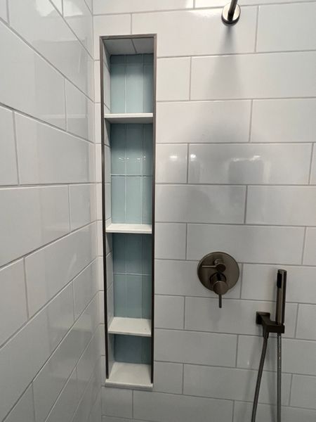 Shower with white tile, a tall built-in shelf with three white shelves and blue tile, and a shower head.