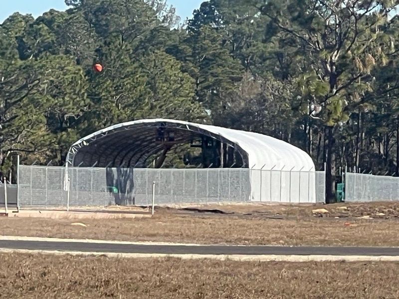 Arched white building behind a chain link fence, set against a backdrop of trees.