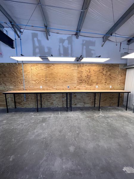 Long wooden workbench against plywood wall under lights in workshop.