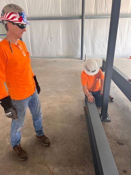 Two construction workers in orange shirts and hard hats, inspecting a metal beam inside a building.