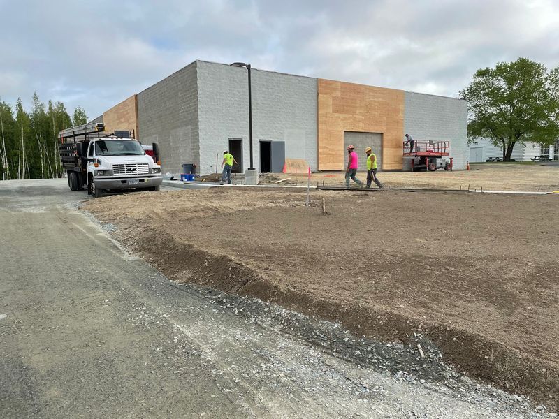 Construction site with a tan brick building, gravel parking area, and small construction equipment. Cloudy sky.