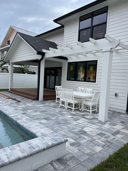 Backyard patio with dining set, pool, and pergola, attached to a white house with black window frames.