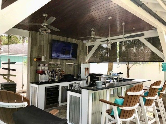 Covered outdoor bar with striped stools, TV, ceiling fans, and white counters on a patio.
