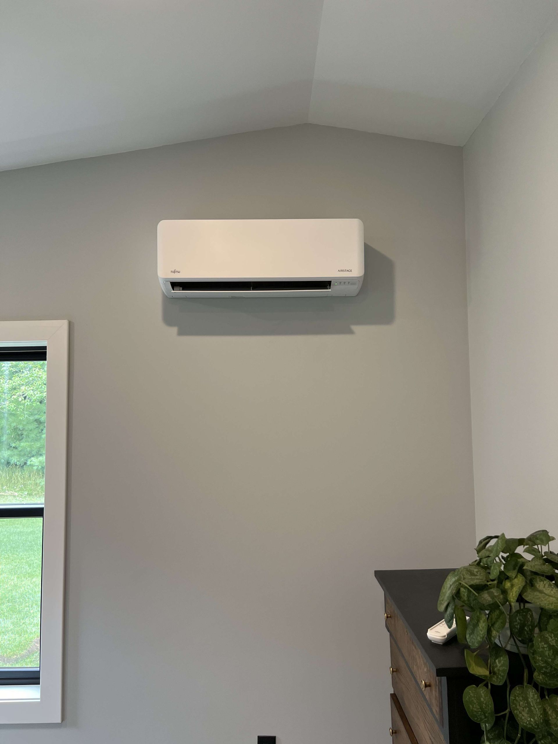 White wall-mounted air conditioner on a gray wall above a window and potted plant.