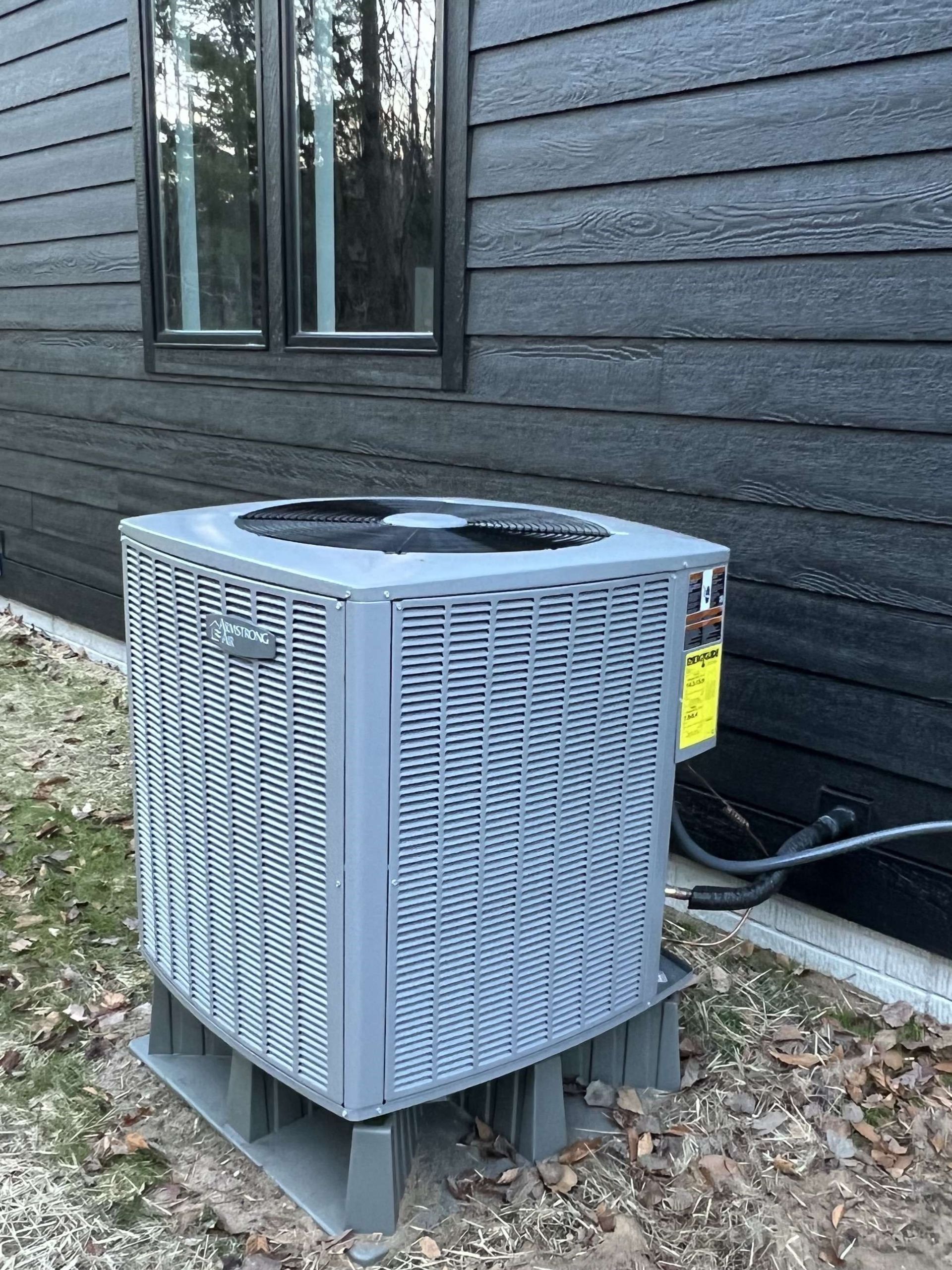 Outdoor air conditioner unit beside a dark gray house wall, sitting on a concrete pad amid leaves.