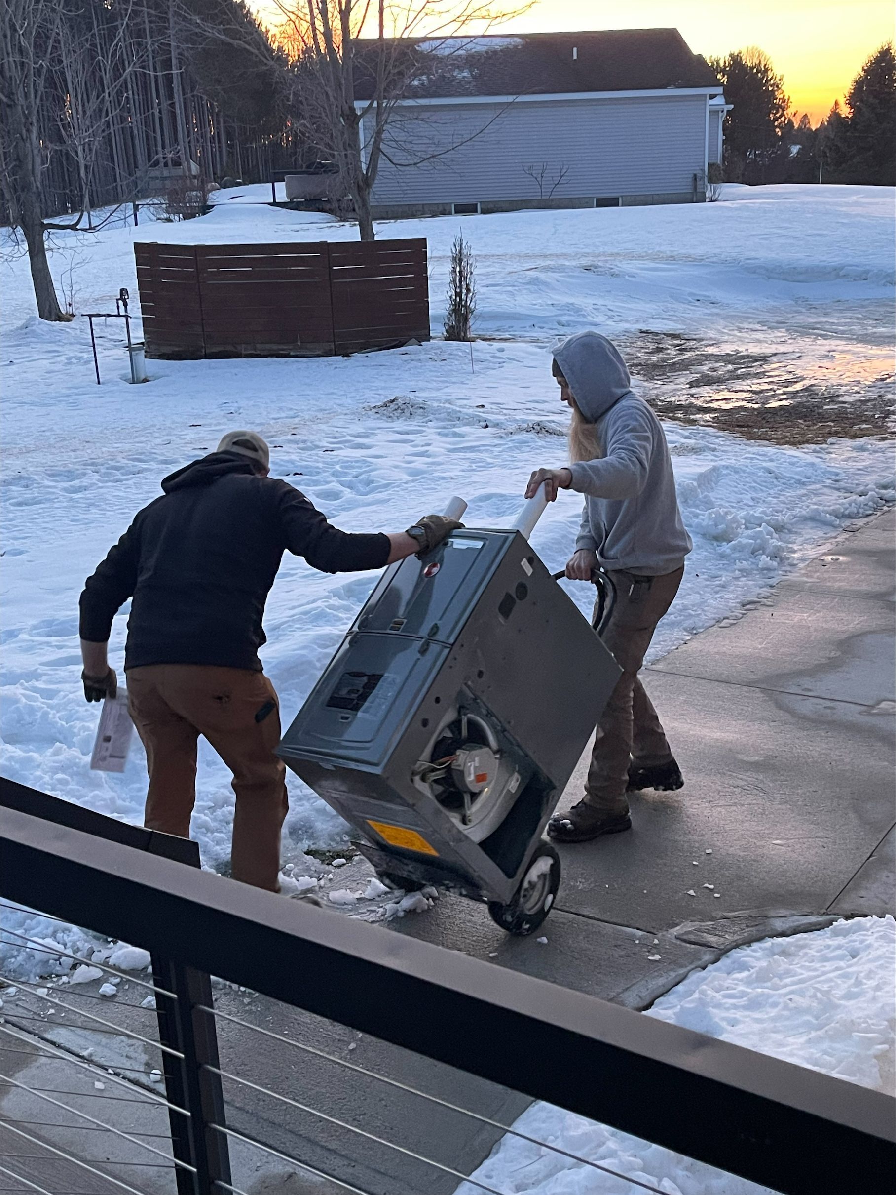 Two people dragging a tipped washing machine across a snowy driveway at dusk