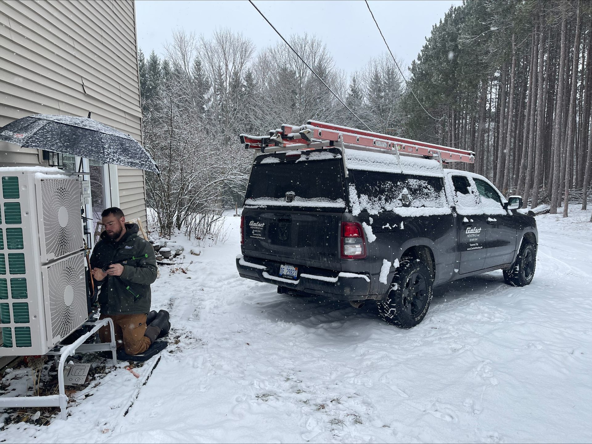 Snow-covered driveway with a black SUV, ladder on top, and a person sitting by the house near outdoor equipment.