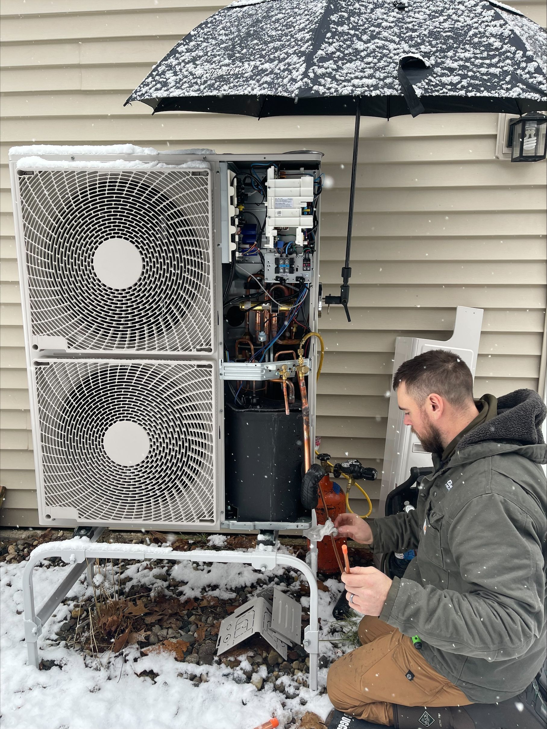 Technician repairing an outdoor HVAC unit in snow beside a house, with equipment panels open.