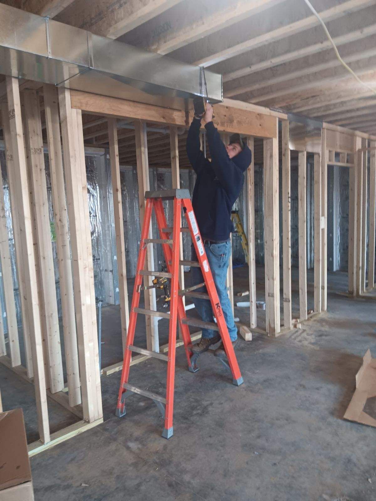 Worker on a red ladder installing overhead framing in an unfinished room.