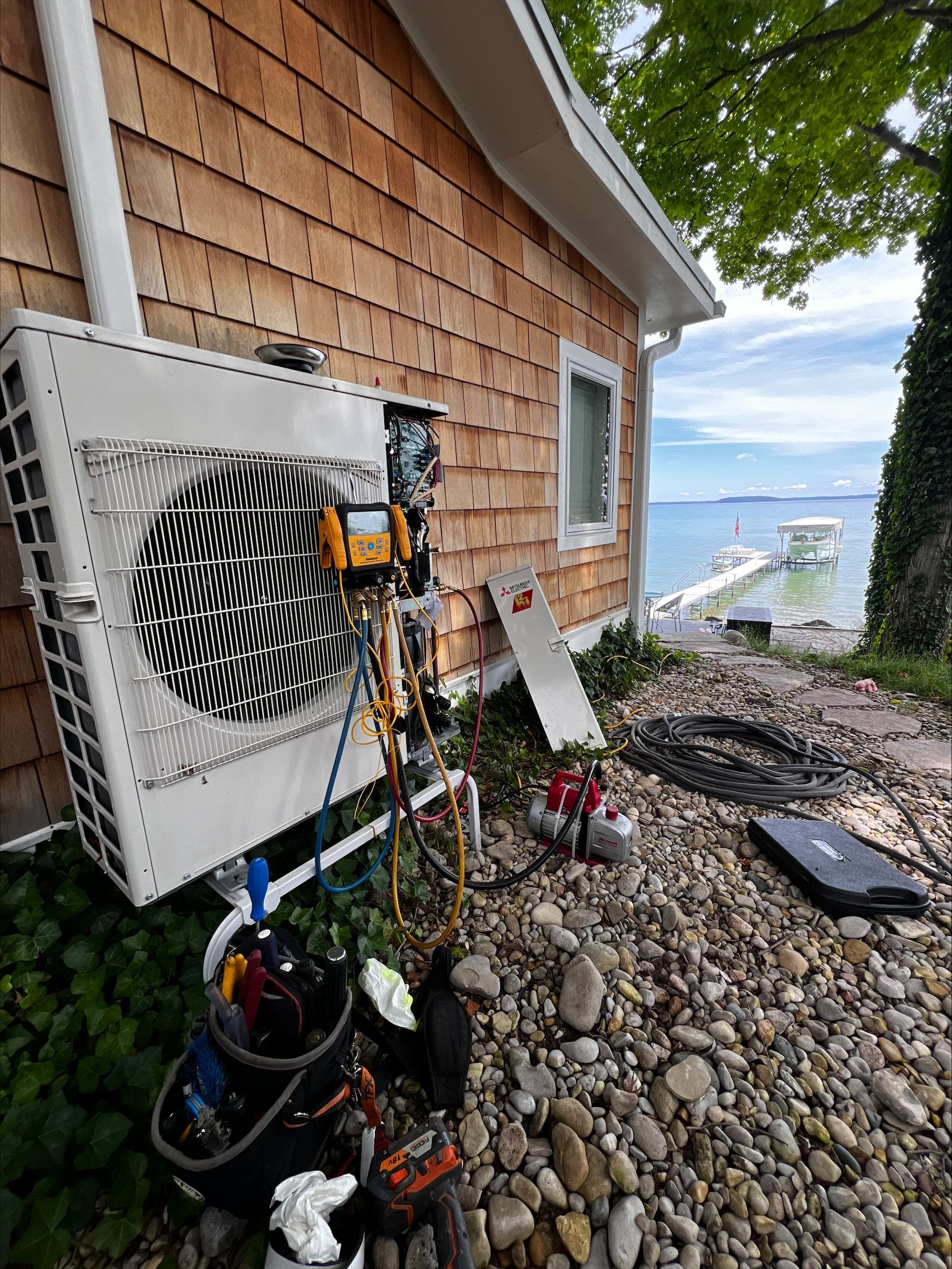 Outdoor heat pump unit mounted beside a lakeside house, with tools and rocks on the ground.