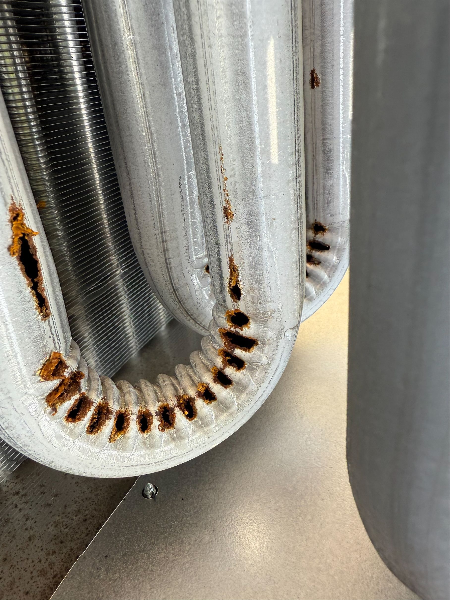 Rust on corrugated metal duct beside a gray cylindrical pipe on a speckled floor