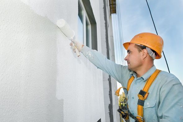 A construction worker in a safety harness paints the exterior of a building with white paint.