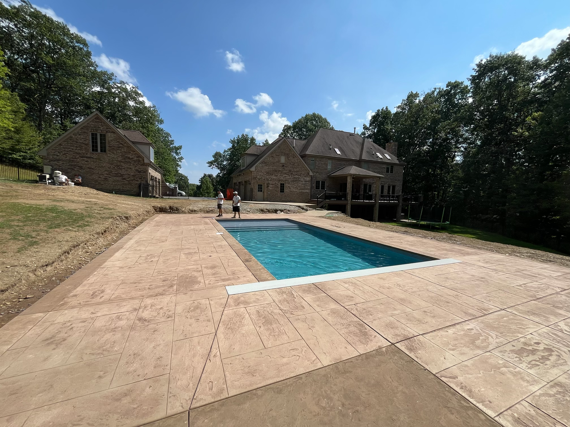 Rectangular pool with blue water, surrounded by tan concrete. Two-story brick house in background under blue sky.
