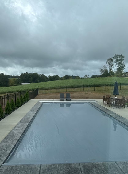 A rectangular pool with dark gray water and patio furniture against a cloudy sky and green field.