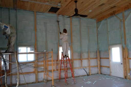 A man is standing on a ladder spraying insulation on a wall.