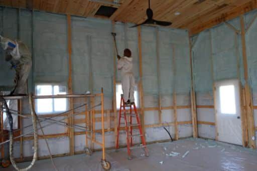 A man is standing on a ladder spraying insulation on a wall.