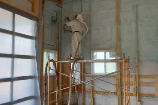 A man is spraying insulation on a wall while standing on a scaffolding.