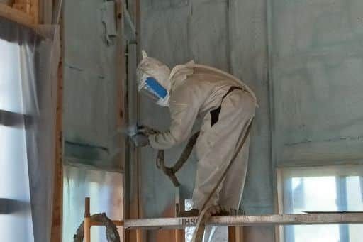 A man in a protective suit is spraying insulation on a wall.