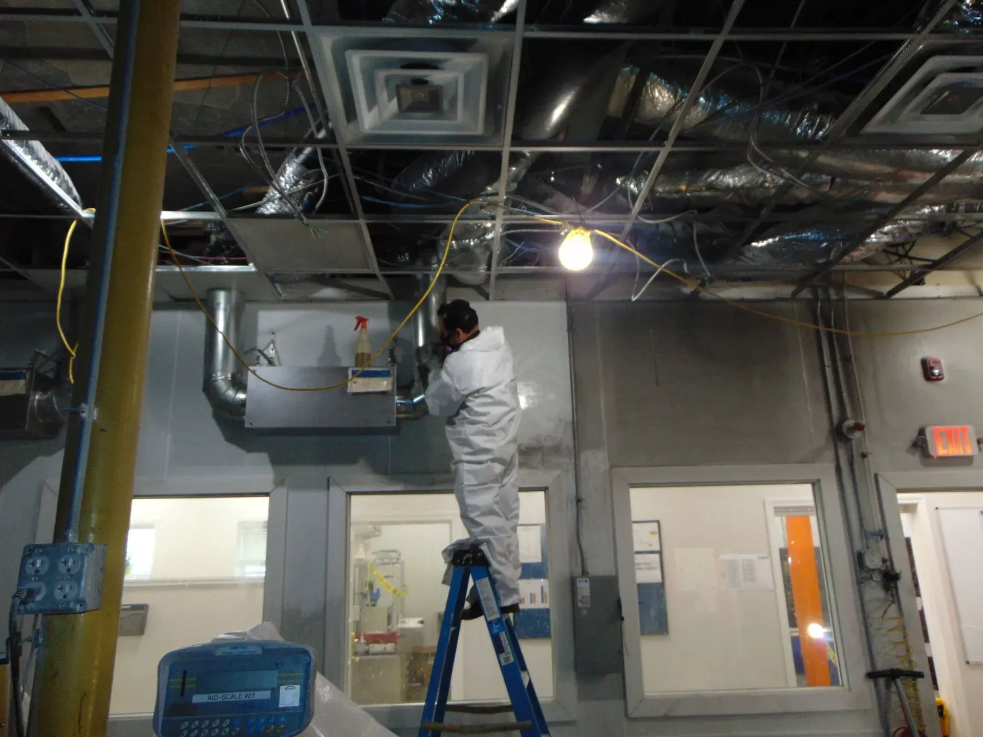 Person in protective suit on ladder, working on electrical wiring in a commercial building.