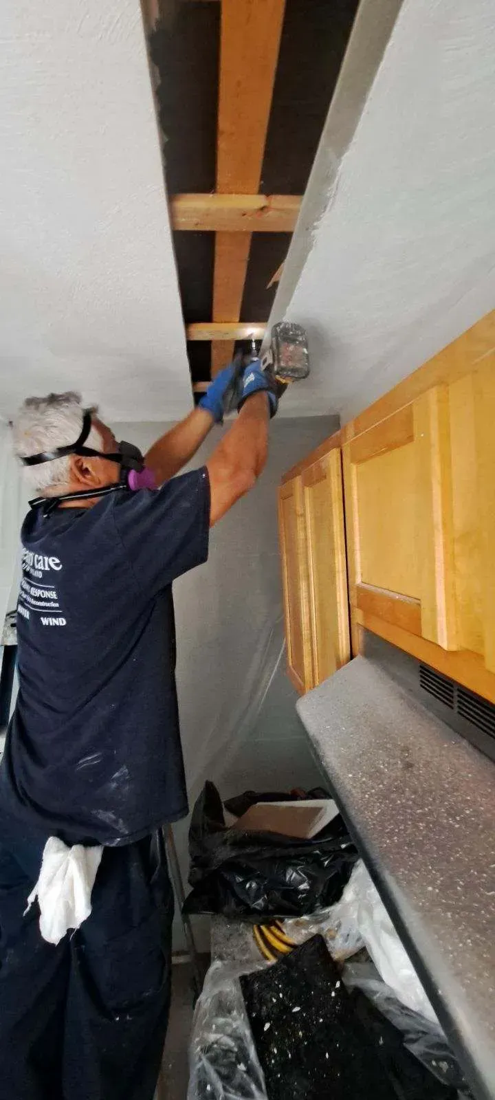 Person in protective gear using a tool to cut ceiling near kitchen cabinets.
