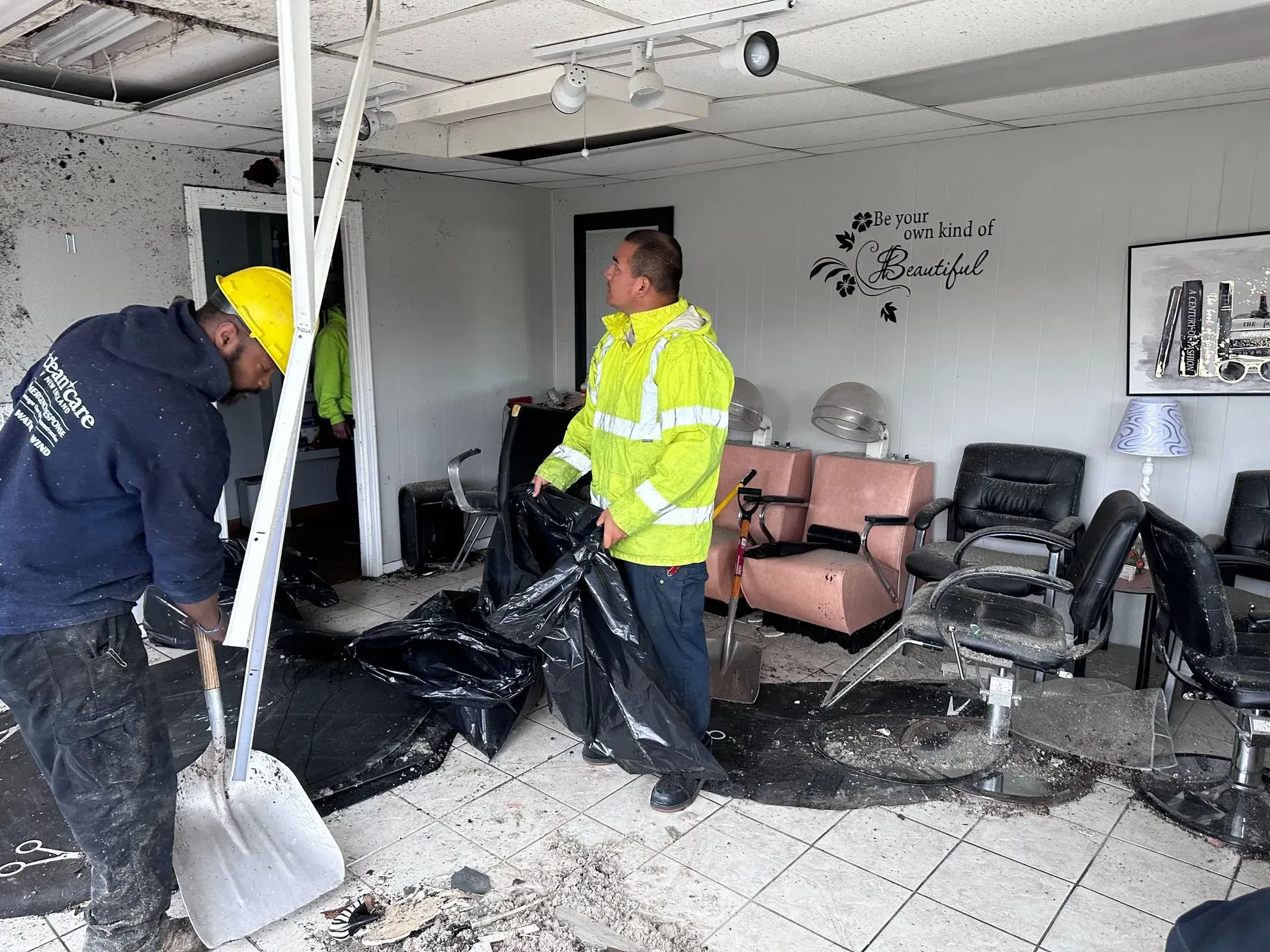 Two people cleaning a damaged salon. One uses a shovel, the other holds a trash bag. Debris litters the floor.