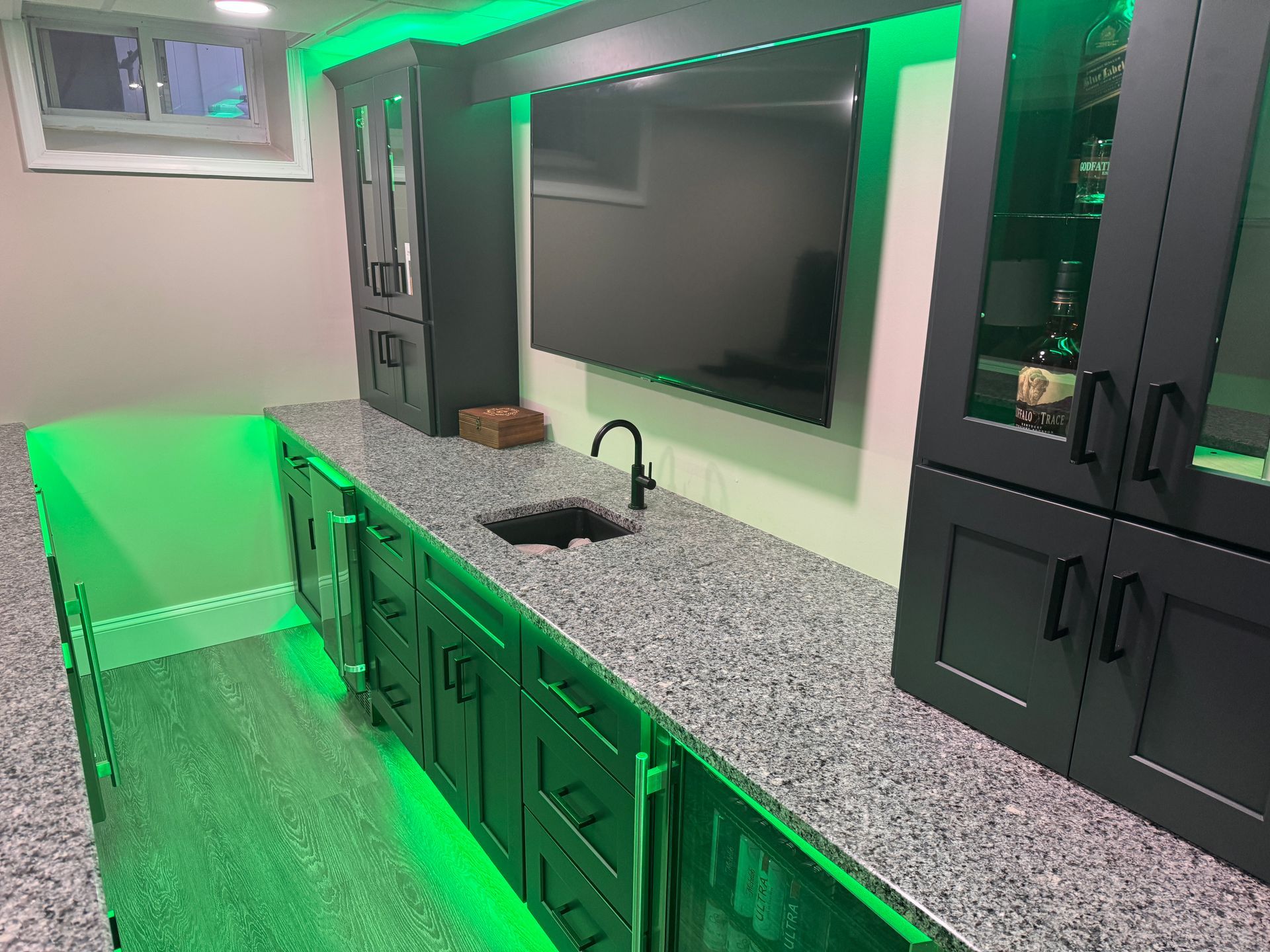 A modern bar area with dark cabinets, green underlighting, and a gray countertop with a sink and a black faucet.