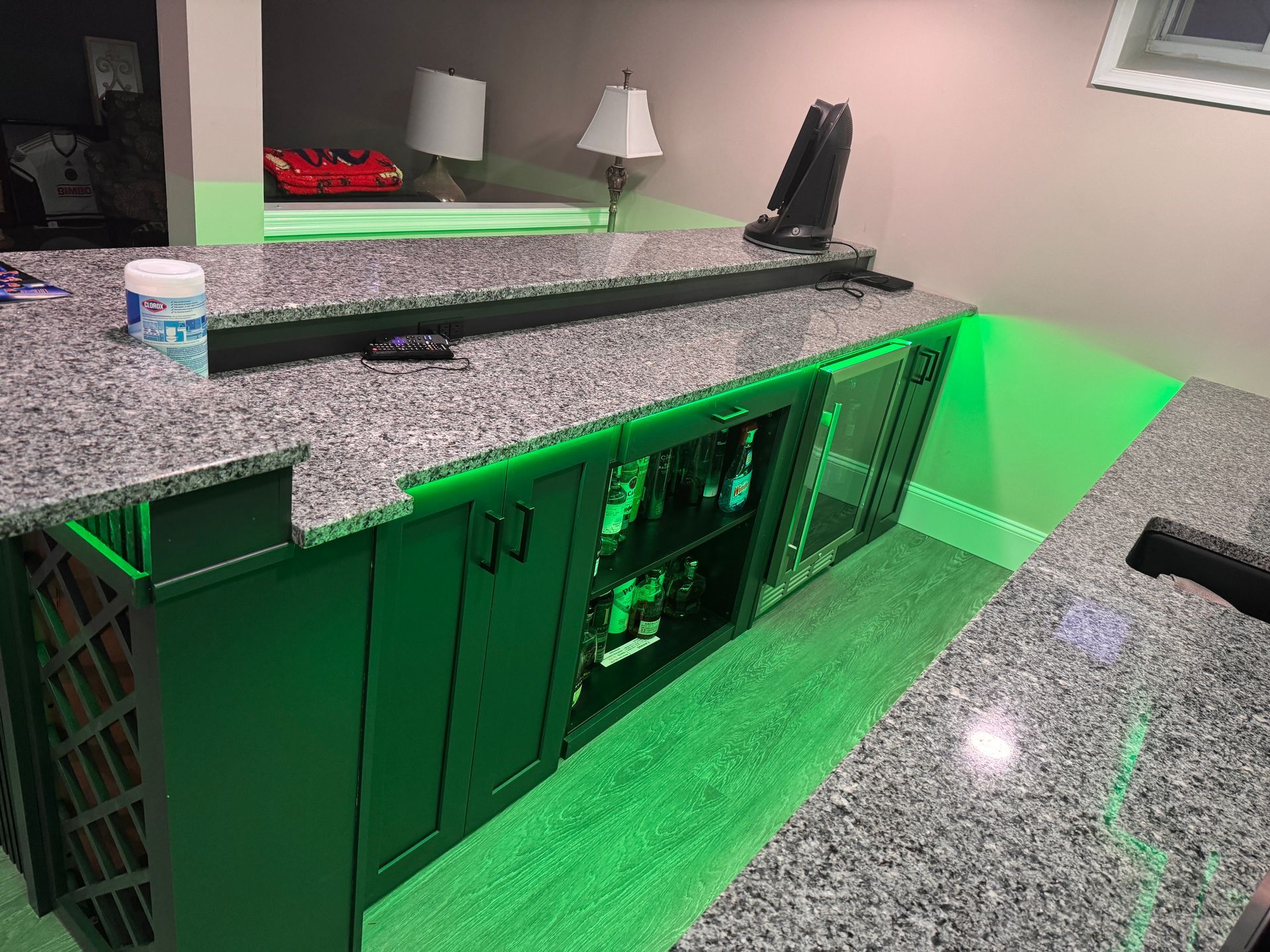 Green-lit bar with granite countertop and cabinets, drinks inside, and a speaker on top.