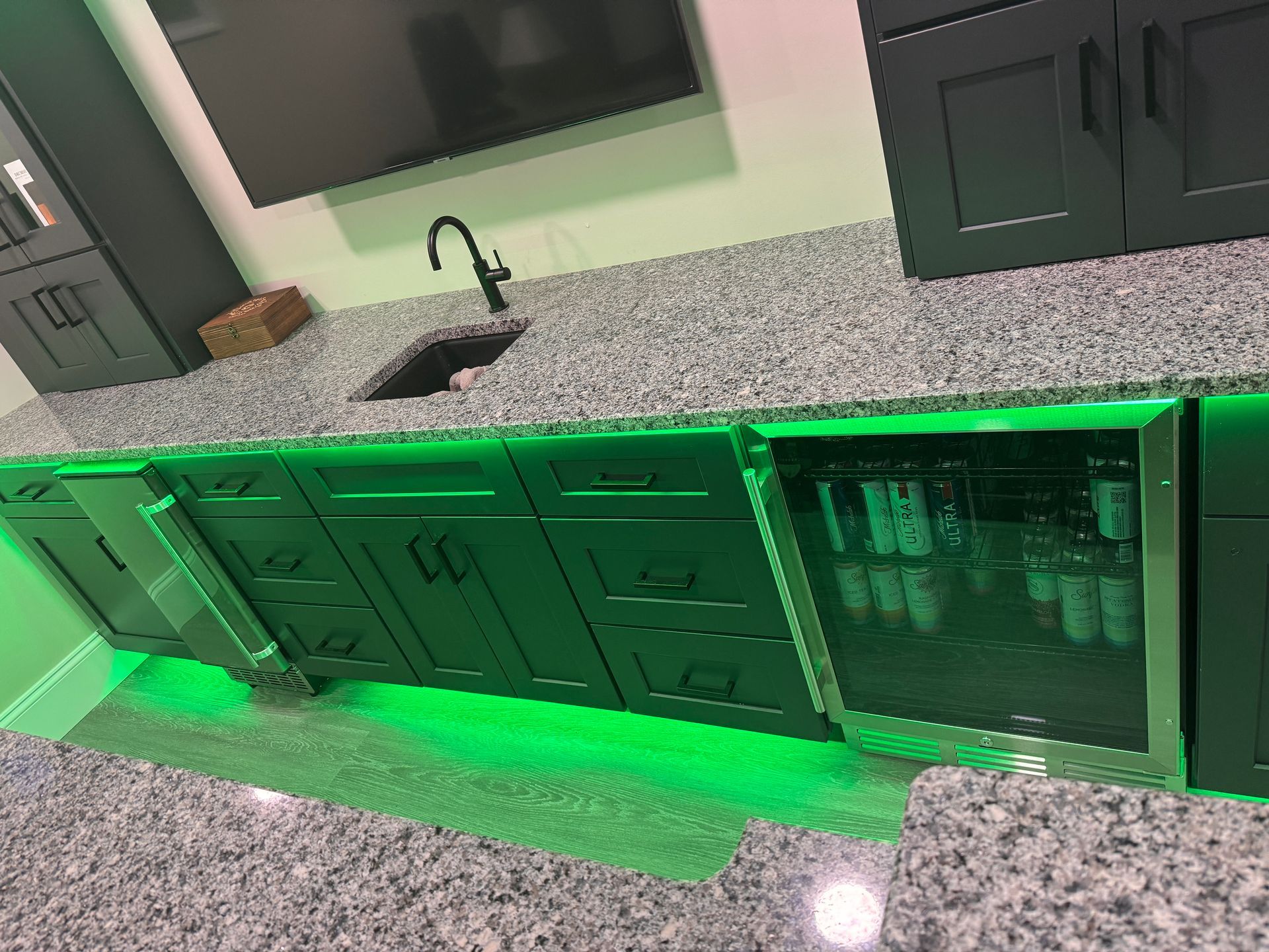 Dark cabinets with granite countertop, sink, and under-cabinet green lighting, alongside a beverage fridge and TV.