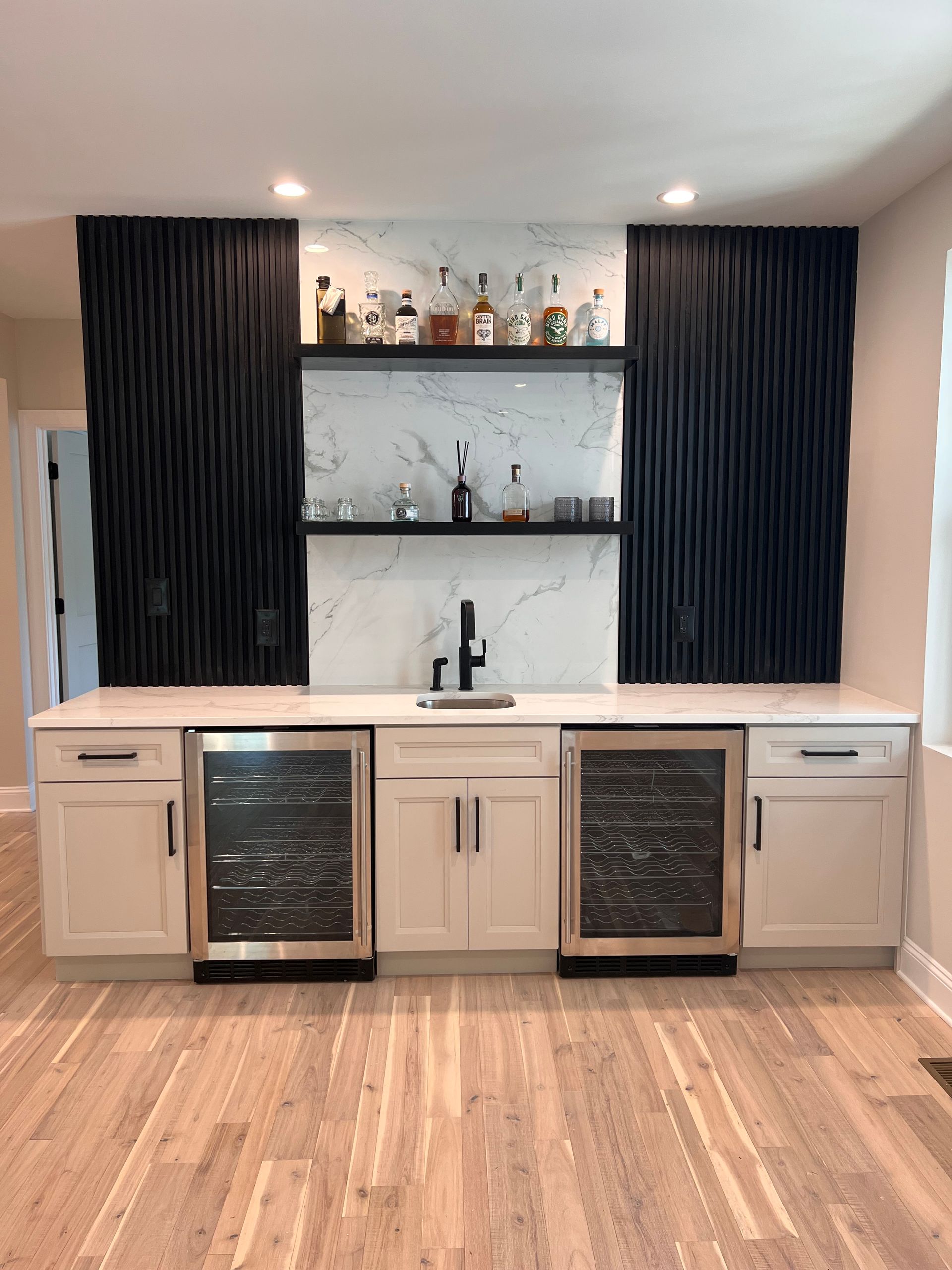 Built-in home bar with black accents and a marble backsplash. Includes white cabinets, wine coolers, and liquor display.