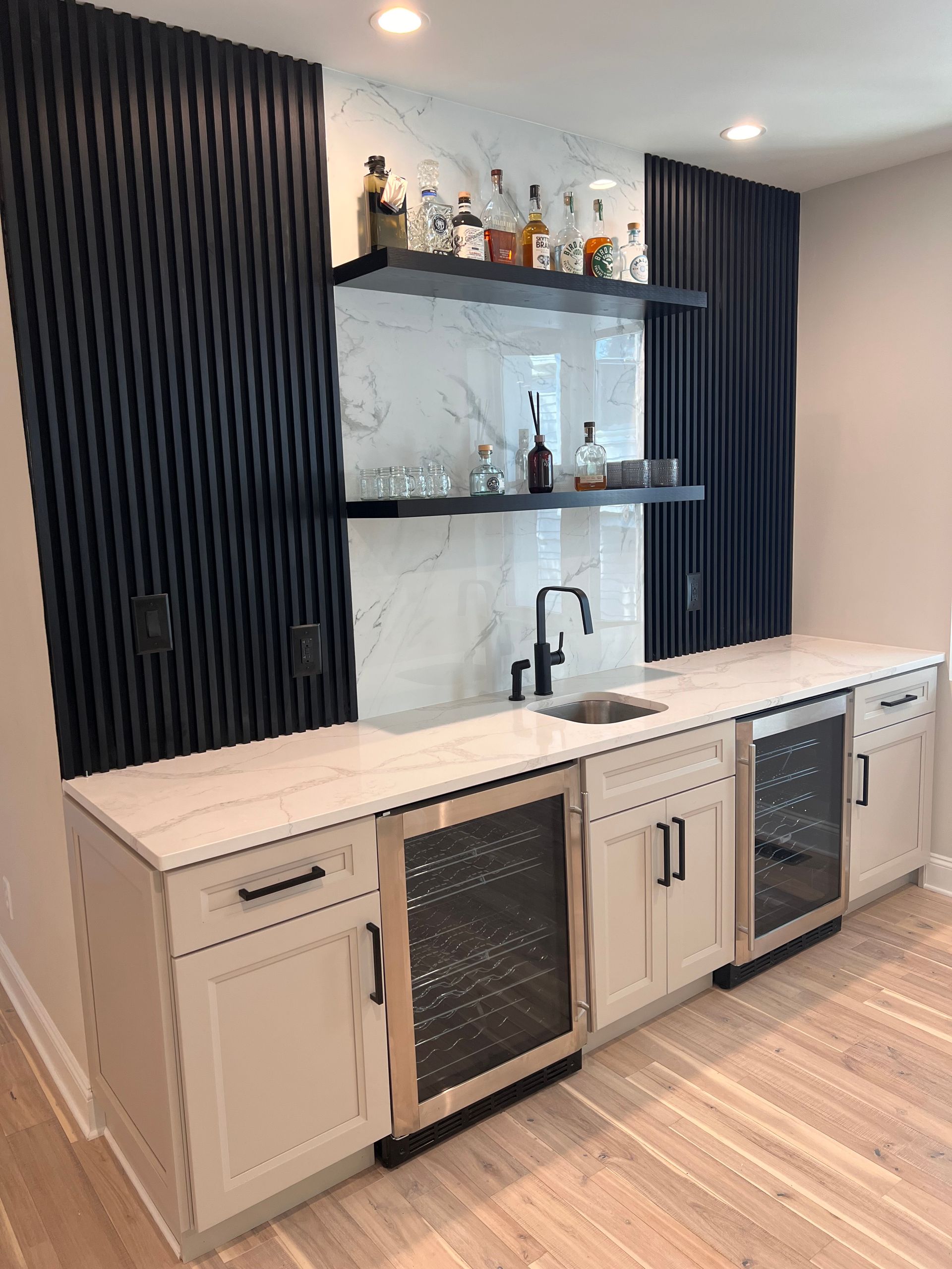 Home bar with black accent wall, white countertop, sink, and cabinets with wine coolers.